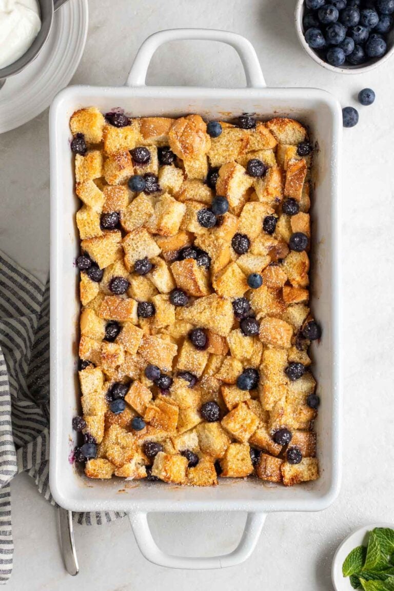 White baking dish of blueberry French toast casserole with powdered sugar sprinkled on top, fresh blueberries, fresh mint, a stripped blue tea towel and silver spatula on the side on the white background.
