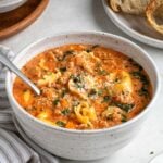 Close up of a white bowl of creamy sausage tortellini soup with a silver spoon with freshly grated parmesan cheese on the side and a plate of crusty bread in the background with a striped white and grey tea towel on a white background.