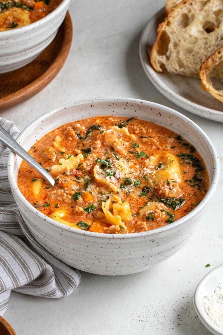Close up of a white bowl of creamy sausage tortellini soup with a silver spoon with freshly grated parmesan cheese on the side and a plate of crusty bread in the background with a striped white and grey tea towel on a white background.