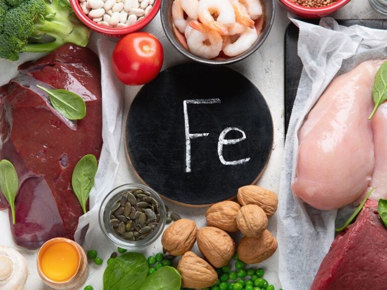 Iron symbol written in chalk on a small round chalkboard surrounded by liver, broccoli, white beans, tomatoes, shrimp, lentils, chicken breasts, red meat, walnuts, green peas, spinach, pumpkin seeds, eggs, and mushrooms.