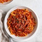 White bowl of lentil bolognese spaghetti with fresh parmesan and chopped parsley on top with a silver fork on the side on a white background.