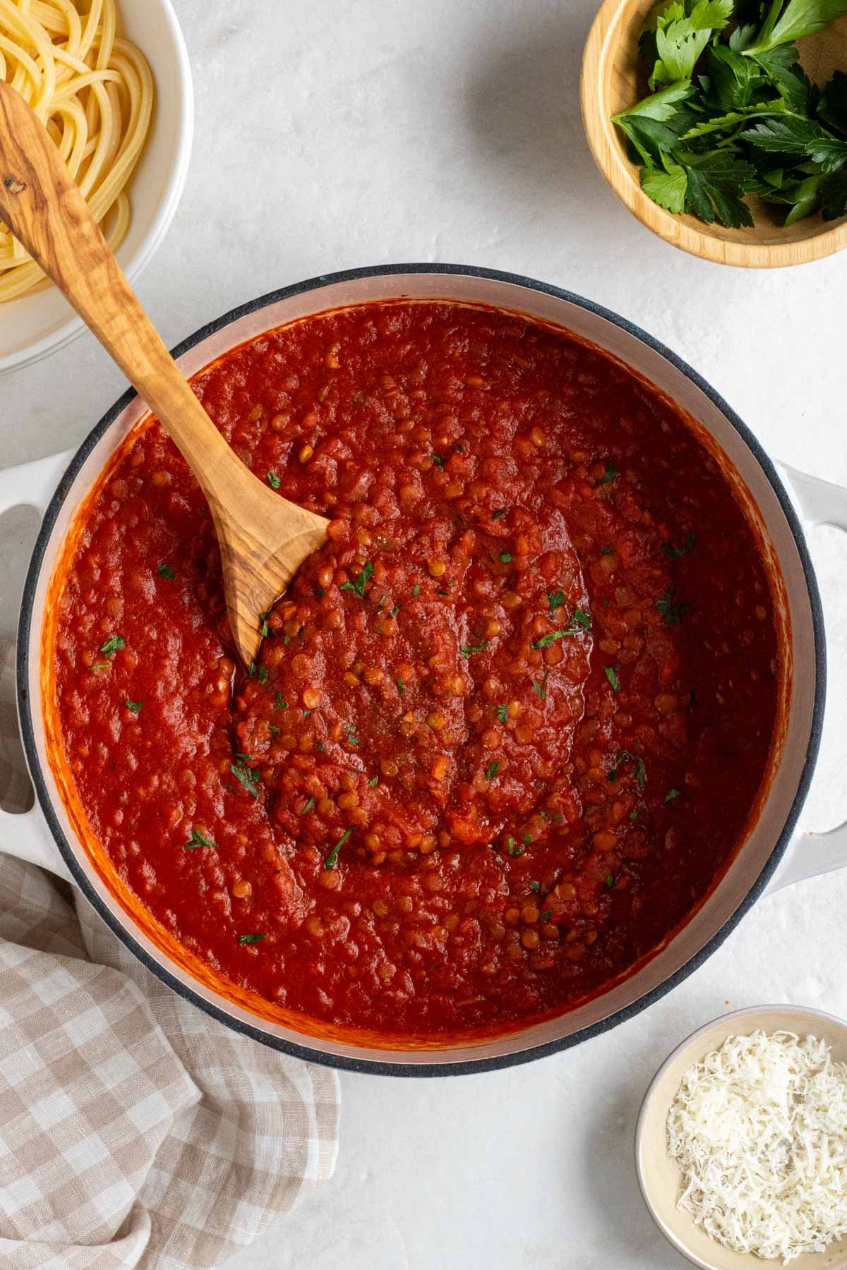 Large pot of lentil bolognese pasta sauce with a wood serving spoon with cooked spaghetti, fresh parsley, freshly grated parmesan cheese, and a beige checkered tea towel on the side on a white background.