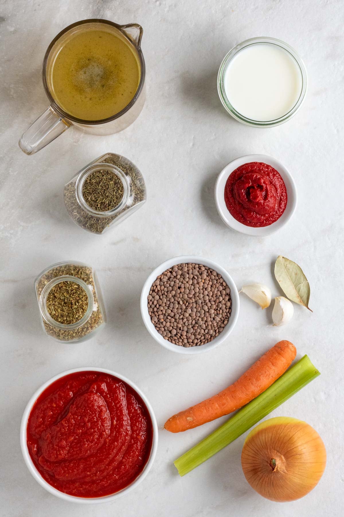 Glass jug of vegetable broth, jar of heavy cream, jars of dried thyme and dried oregano, bowls of tomato paste, brown lentils, and crushed tomatoes with bay leaf, garlic cloves, carrot, celery, and onion on a white background.