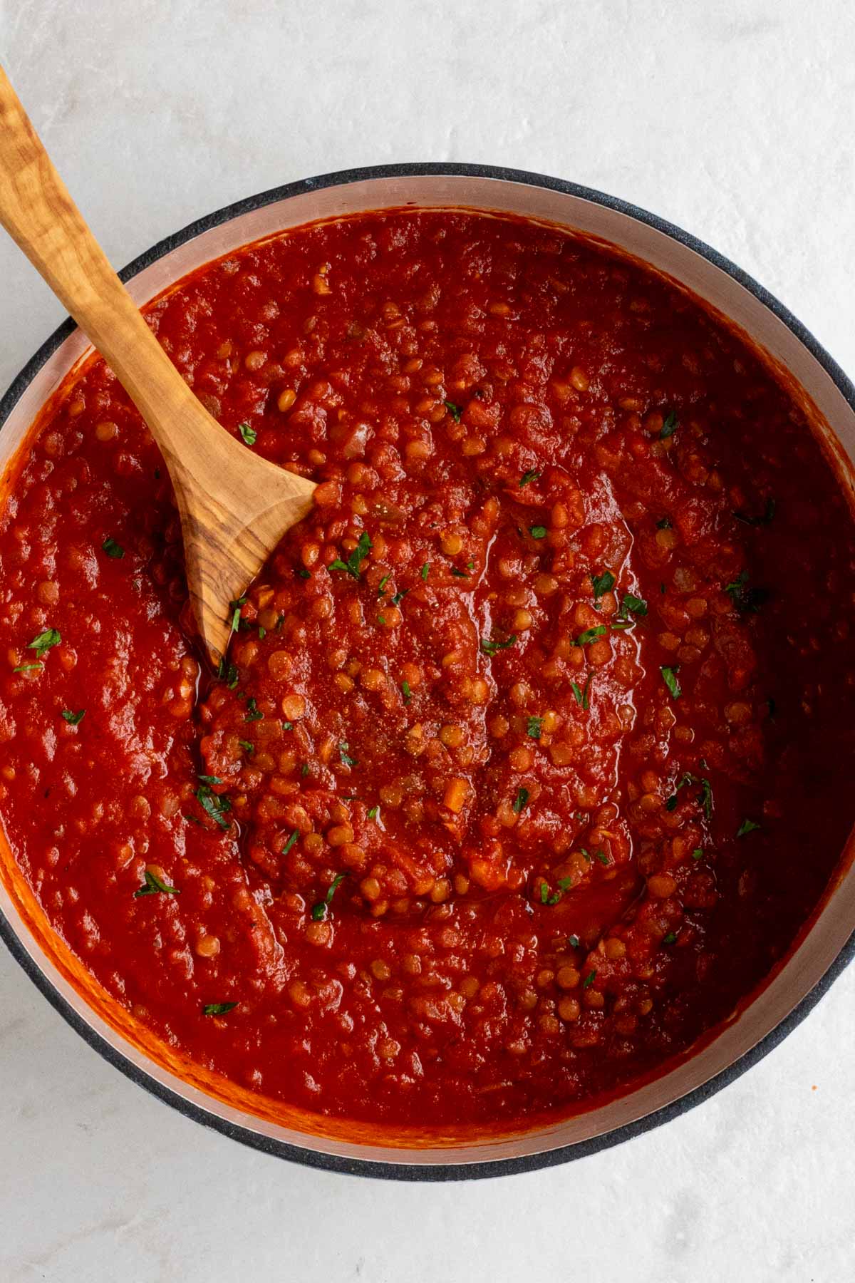 Close up of a bot of lentil bolognese sauce with a wooden spoon on a white background.