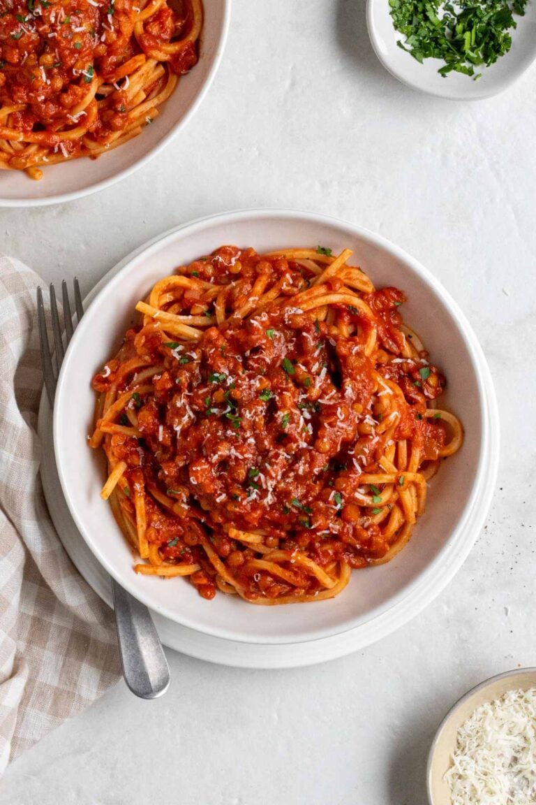 White bowl of lentil bolognese spaghetti with fresh parmesan and chopped parsley on top with a silver fork on the side on a white background.