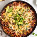 Tex-Mex ground beef skillet with a gold serving spoon with diced avocado, sliced green onion, and lime wedges on top with tortilla chips, salsa, limes, and fresh cilantro on the side with a beige checkered tea towel on a white background.