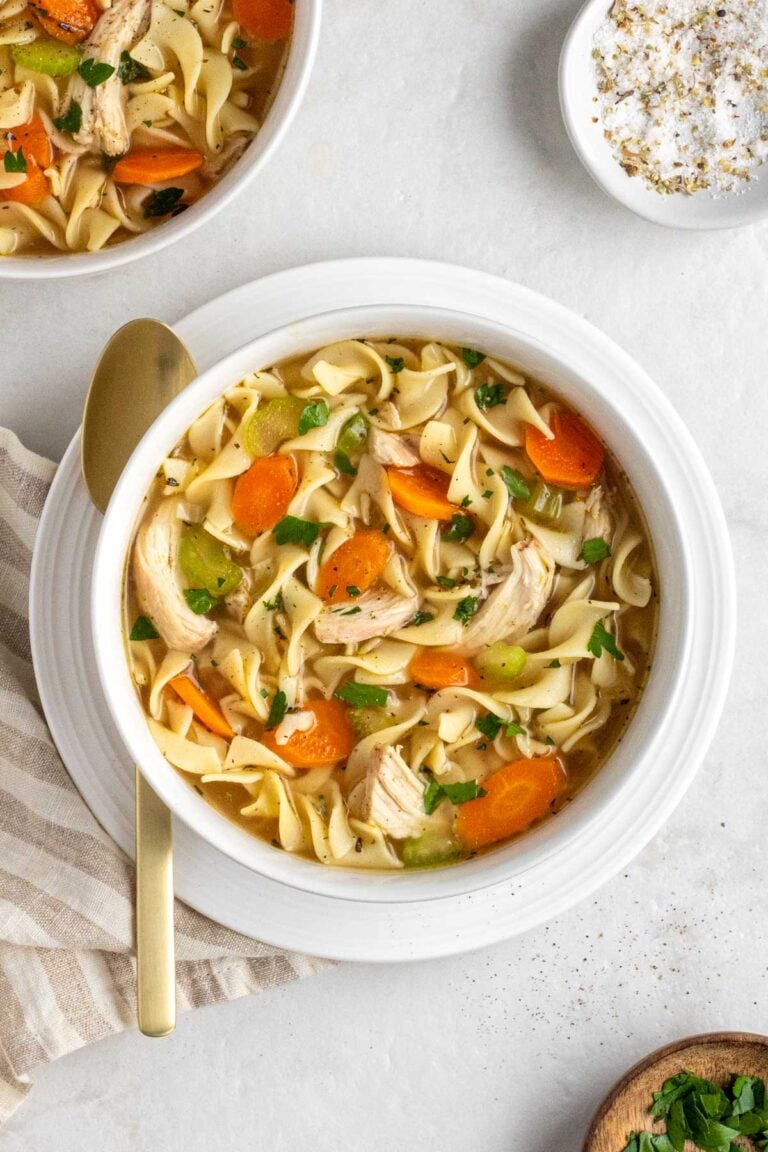 Close up of a white bowl of turkey noodle soup on a small white plate with a gold spoon, beige striped tea towel, white bowl of salt, and small wood bowl of chopped parsley on a white background.