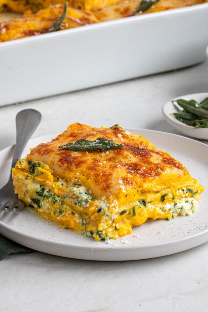 Slice of butternut squash and spinach lasagna on a white plate with freshly grated parmesan cheese and fried sage on top on a white background.