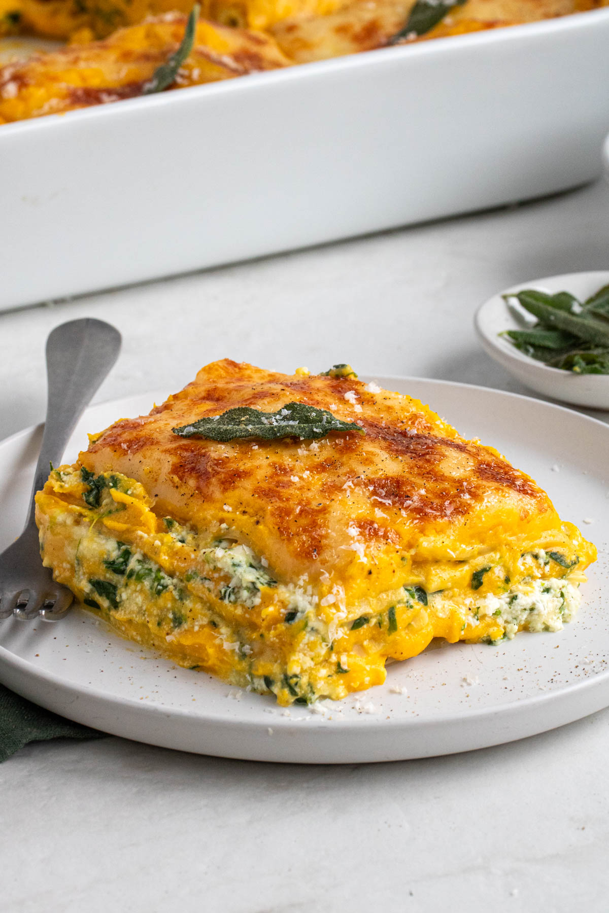 Slice of butternut squash and spinach lasagna on a white plate with freshly grated parmesan cheese and fried sage on top on a white background.