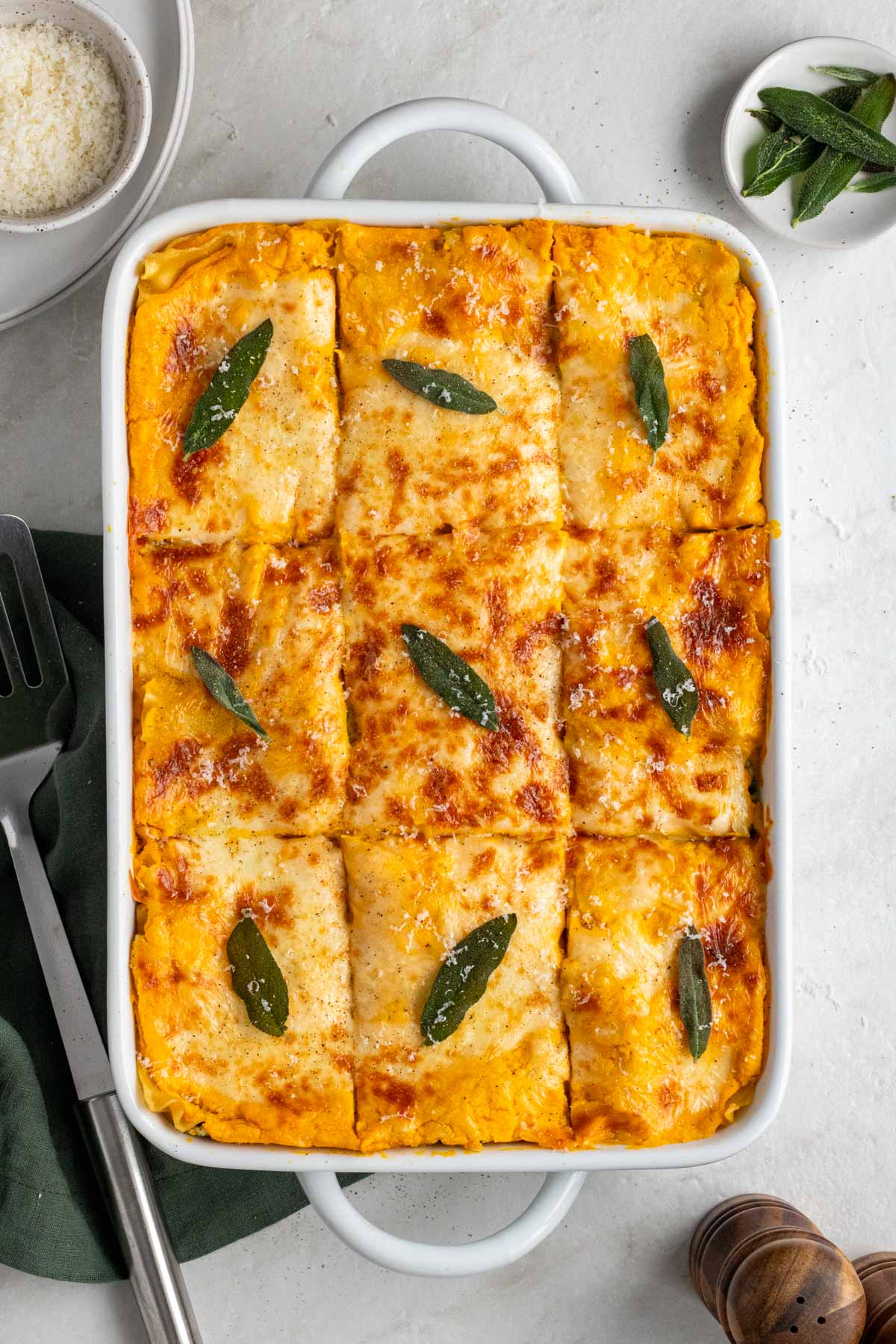 Casserole dish filled spinach and butternut squash lasagna with fried sage leaves and freshly grated parmesan cheese on top with a green tea towel and silver spatula on the side on a white background.