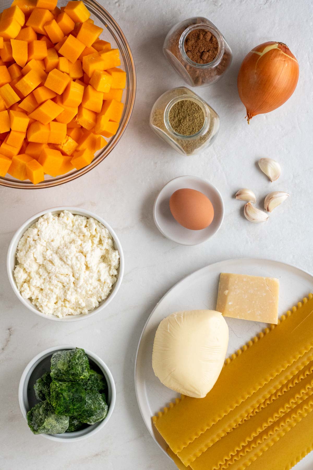 Bowl of cubed ricotta cheese, bowl of frozen spinach with jars of nutmeg and sage, an onion and garlic loves, a small bowl with an egg, and a plate with lasagna noodles, ball of mozzarella cheese, and parmesan cheese on a white background.