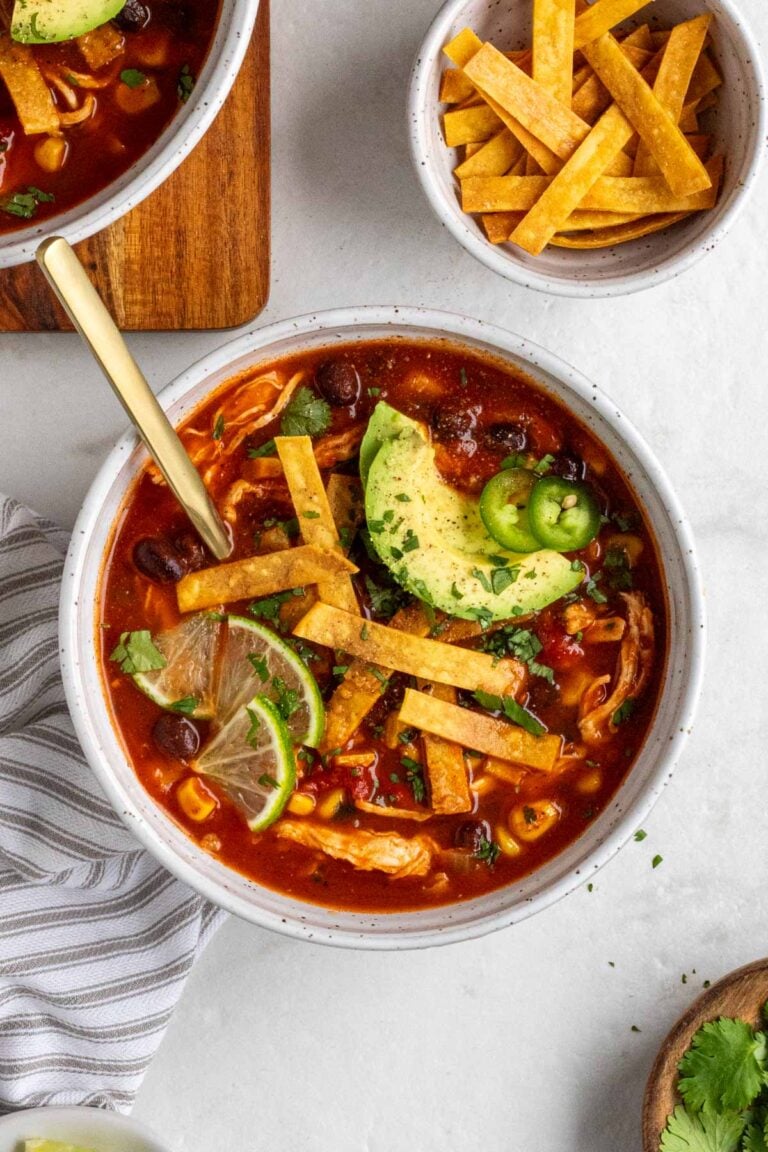 Two bowls of chicken tortilla soup with a gold spoon with a sliced avocado, lime wedges, and fresh cilantro on top and crispy tortilla strips on the side with a grey and white tea towel on a white background.