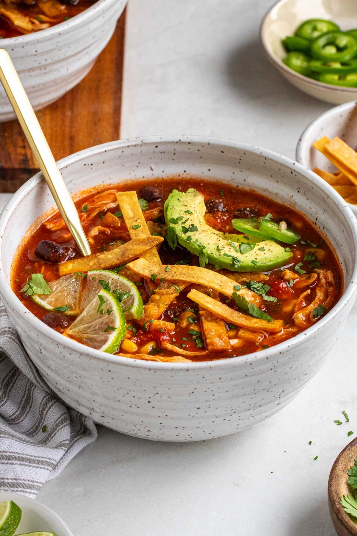 Close up of a speckled white bowl of chicken tortilla soup crushed tortilla strips, lime, avocado, and cilantro on top with a gold spoon on a white background.