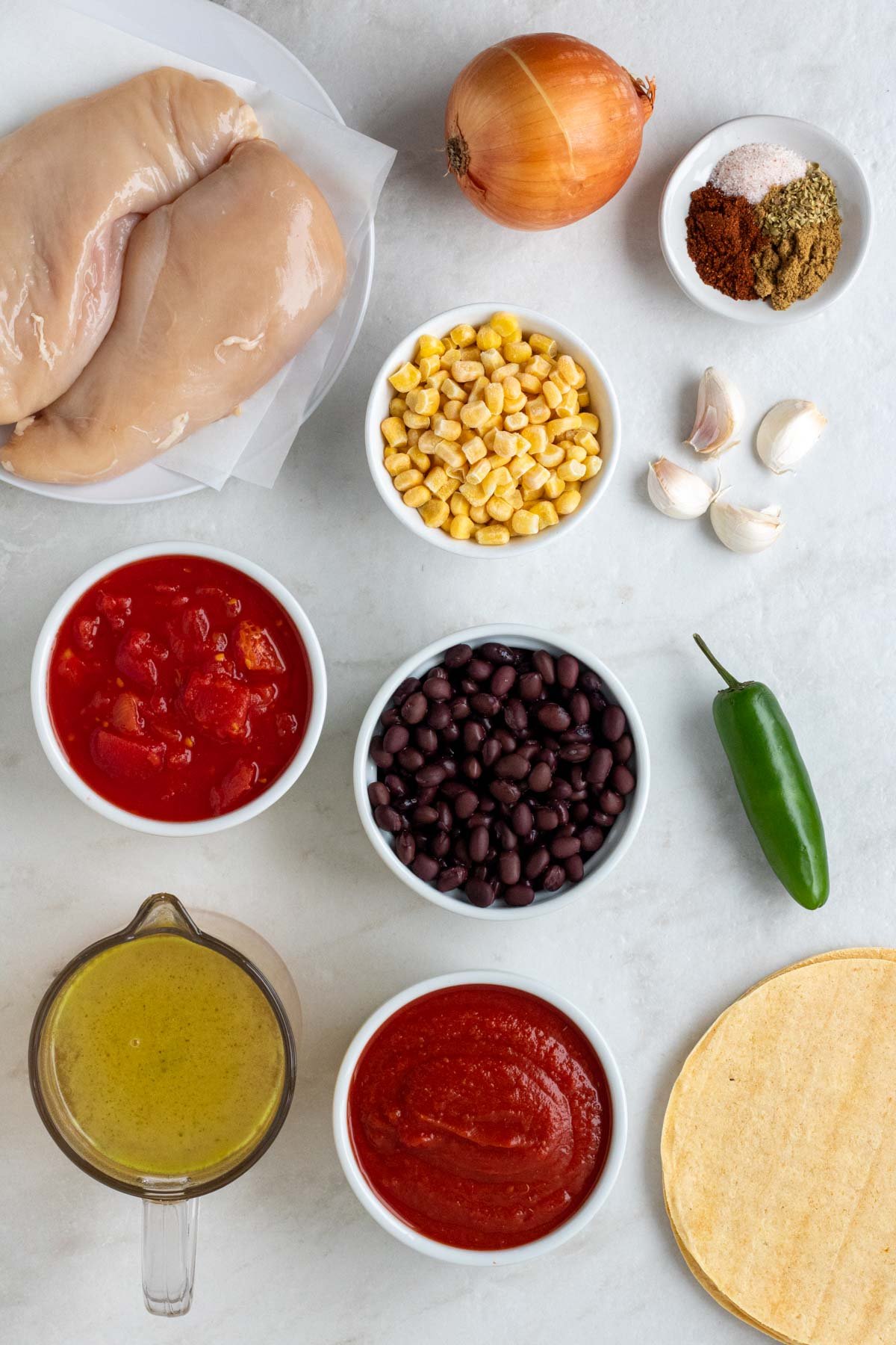 Plate of raw chicken breasts with an onion, jalapeno, garlic cloves, corn tortillas and bowls of spices, frozen corn, black beans, diced tomatoes, crushed tomatoes, and broth.