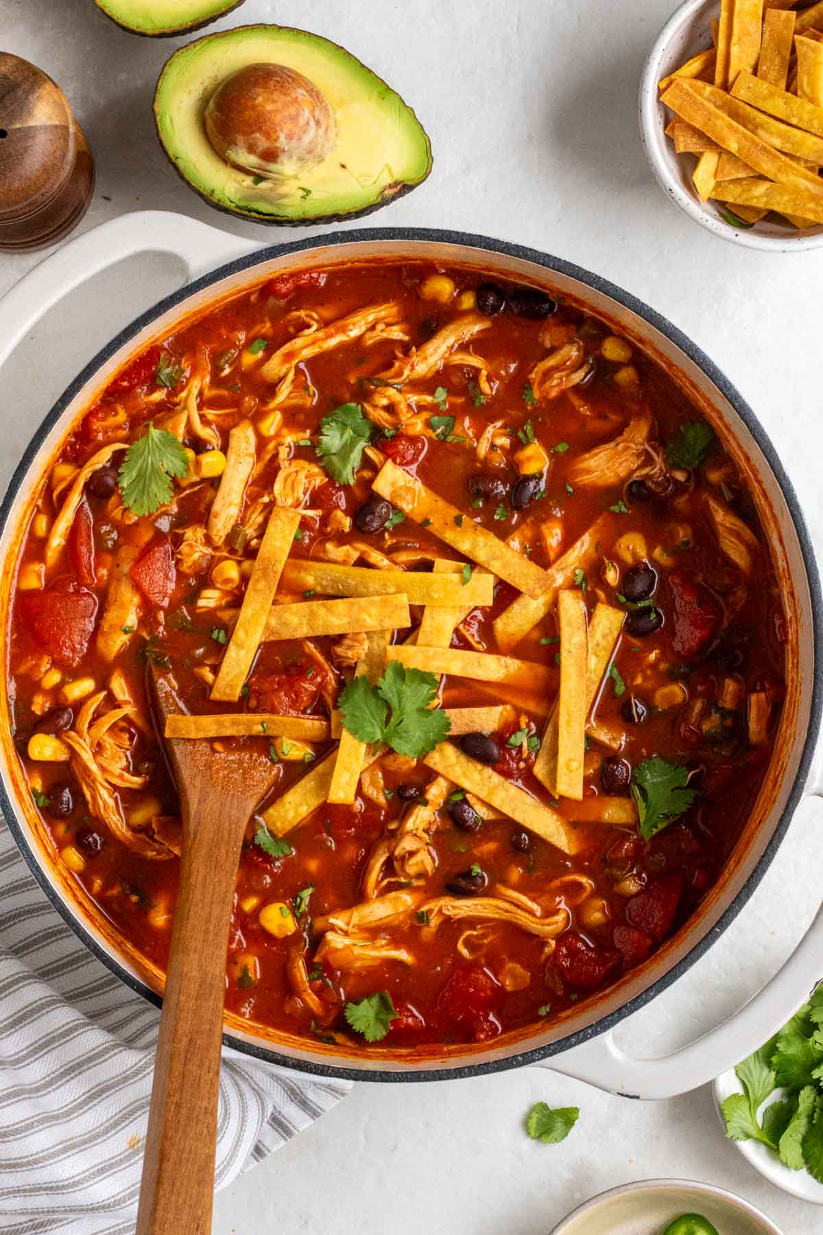 Large pot of chicken tortilla soup with a wooden spoon on a white background with salt, pepper, avocados, a bowl of crispy tortilla strips, and bowl of fresh cilantro on the side with a grey and white tea towel.