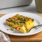Slice of cottage cheese egg casserole on a white plate with a fork on a wood cutting board on a white background.