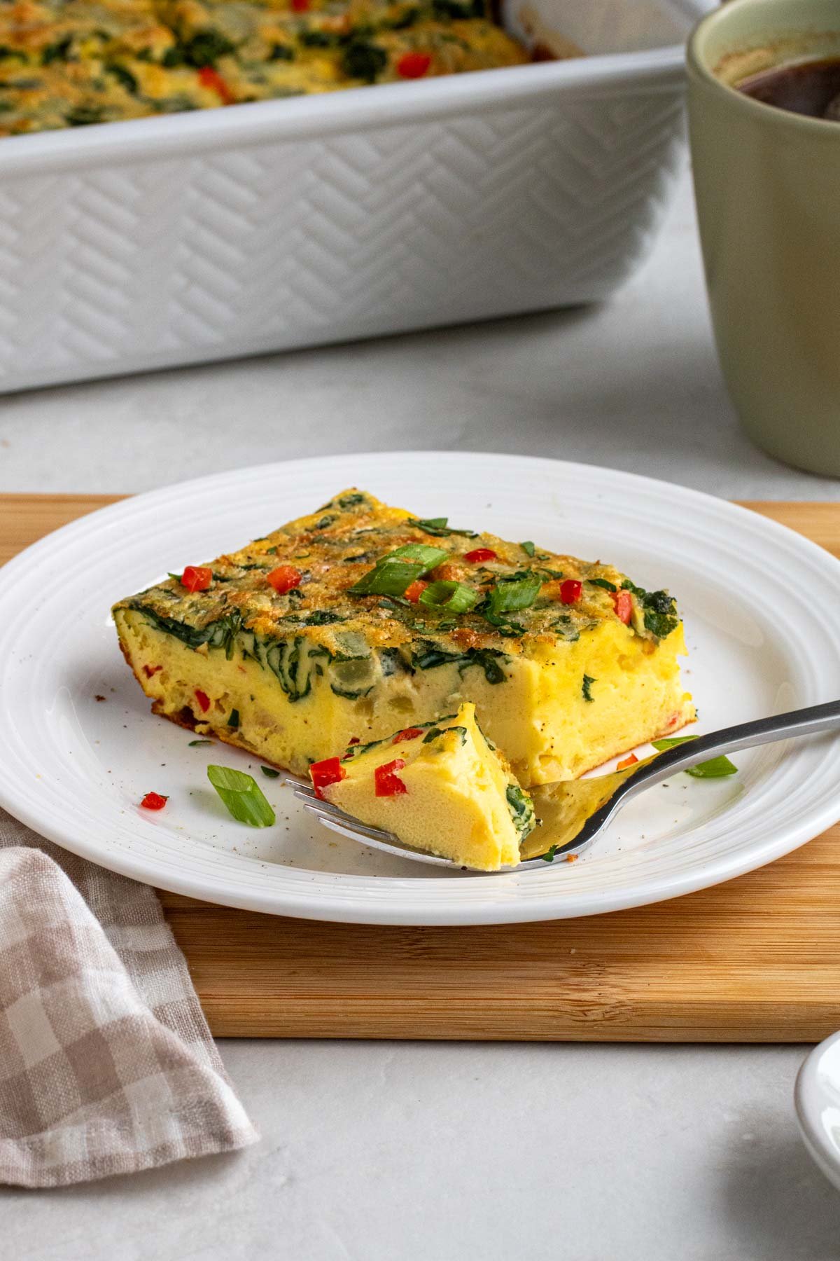Slice of cottage cheese egg casserole on a white plate with a fork on a wood cutting board on a white background.