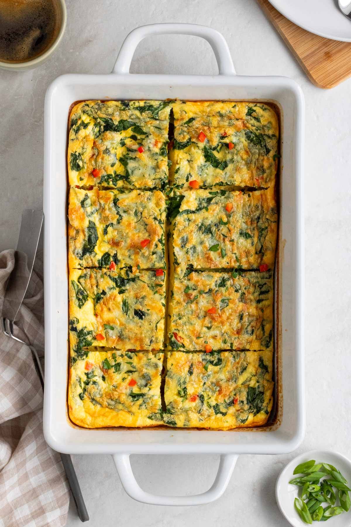 Baking dish of cottage cheese egg bake with a cup of coffee in a green mug and a bowl of sliced green onions and a tea towel on the side on a white background.