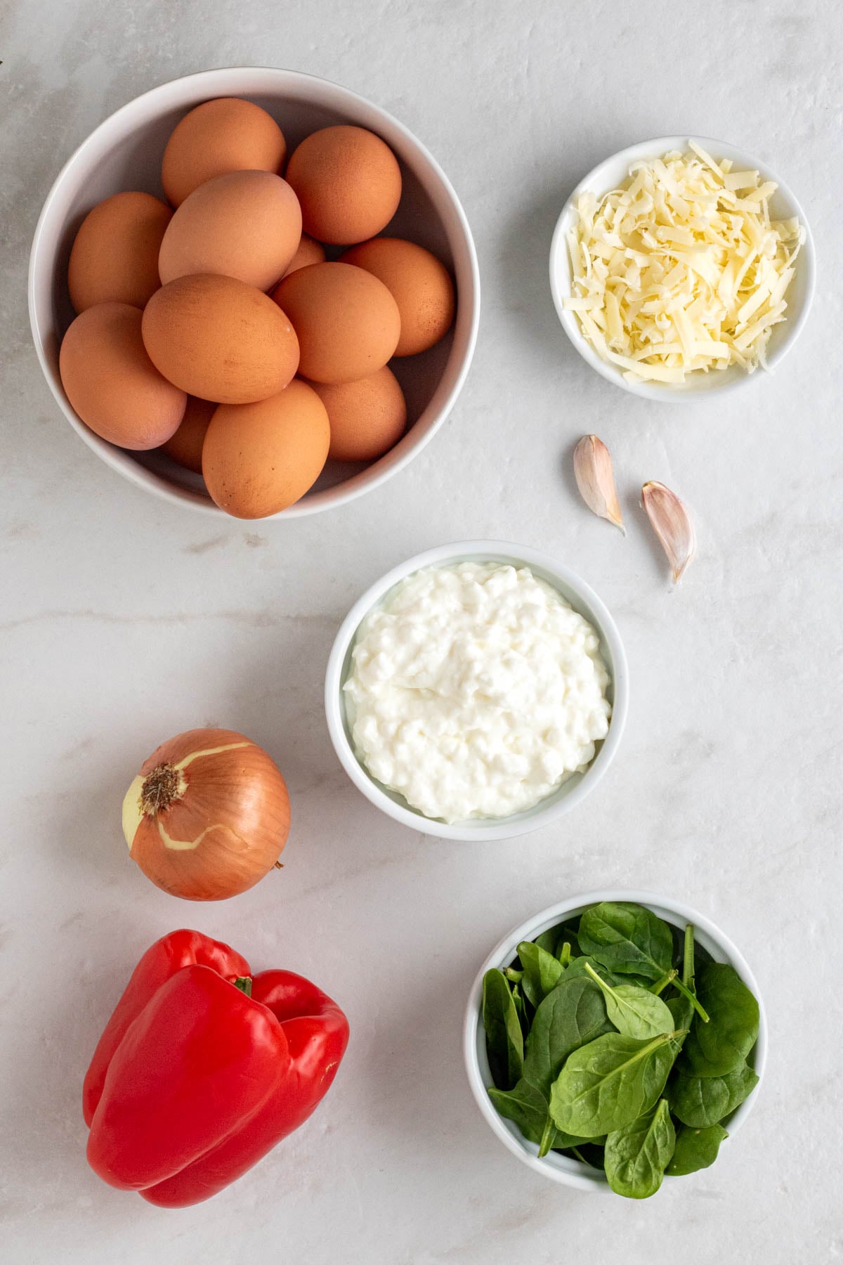 Bowl of eggs, shredded cheese, cottage cheese, and spinach with garlic cloves, onion, and red bell pepper on a white background.