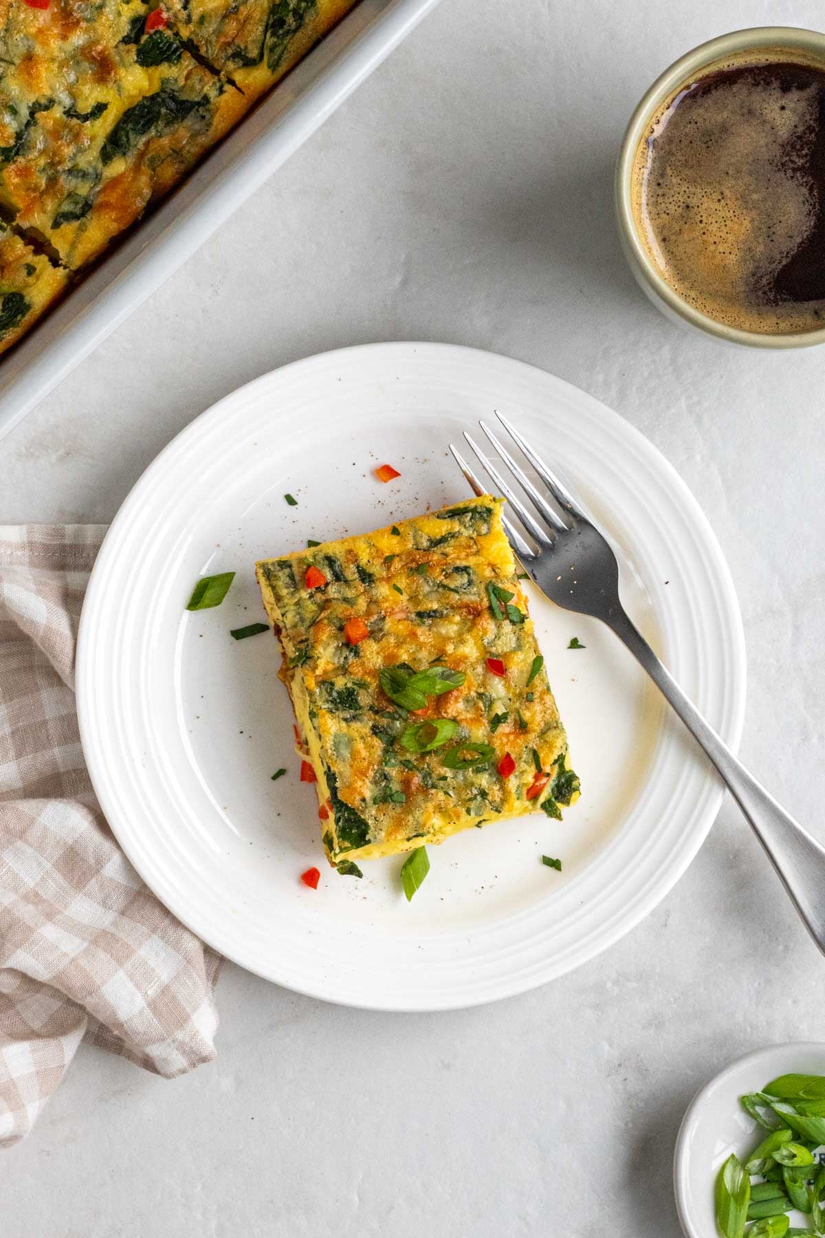 White plate with a slice of cottage cheese egg casserole with a fork and tea towel on the side on a white background.