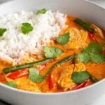 Close up of a bowl of red Thai chicken curry with white rice on the side and fresh cilantro on top with a green tea towel on a white background.