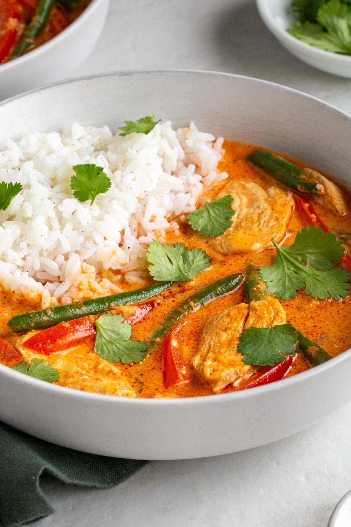 Close up of a bowl of red Thai chicken curry with white rice on the side and fresh cilantro on top with a green tea towel on a white background.