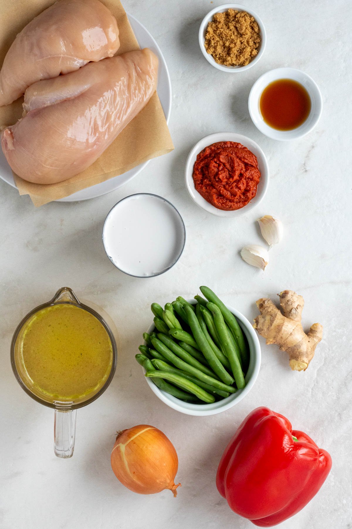 Plate of white chicken breasts with a bowl of brown sugar, fish sauce, and red curry paste with a can of coconut milk with garlic cloves, fresh ginger, green beans, red bell pepper, onion, and broth on a white background.