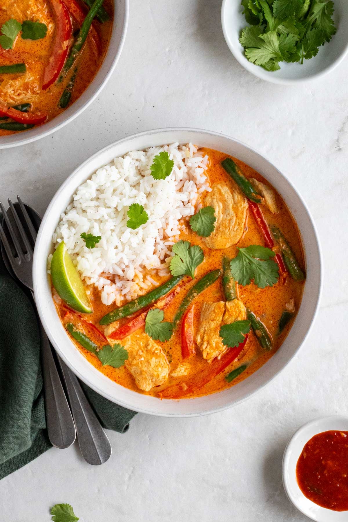 Overhead of two bowls of Thai red chicken curry with white rice and fresh cilantro with a green tea towel, silver spoon and fork, fresh cilantro, and hot sauce on the side.