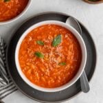 White of of tomato orzo soup with freshly grated parmesan cheese and fresh basil on top on a dark grey plate with a silver spoon with a striped grey and white tea towel, wood bowl of basil leaves, plate of sliced bread, and bowl of black pepper on the side on a white background.