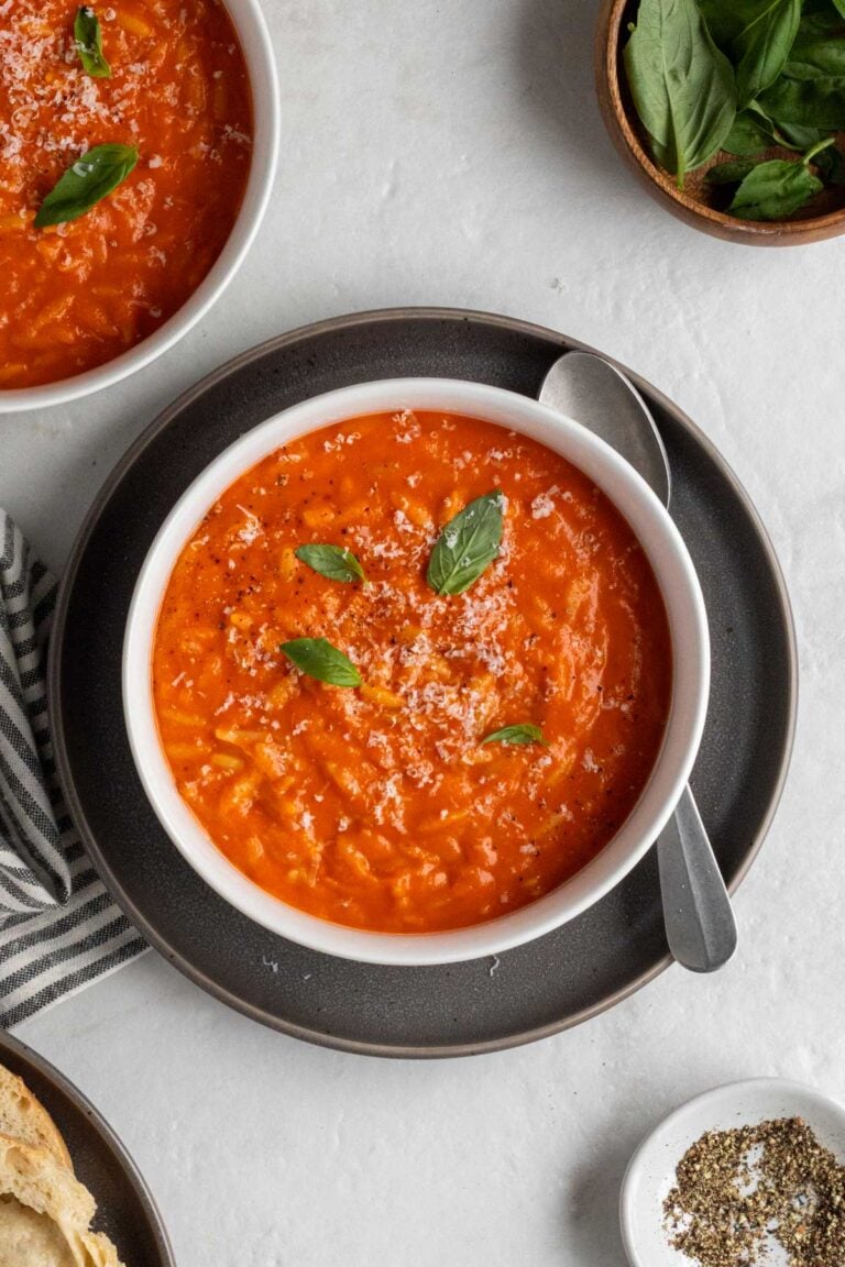White of of tomato orzo soup with freshly grated parmesan cheese and fresh basil on top on a dark grey plate with a silver spoon with a striped grey and white tea towel, wood bowl of basil leaves, plate of sliced bread, and bowl of black pepper on the side on a white background.