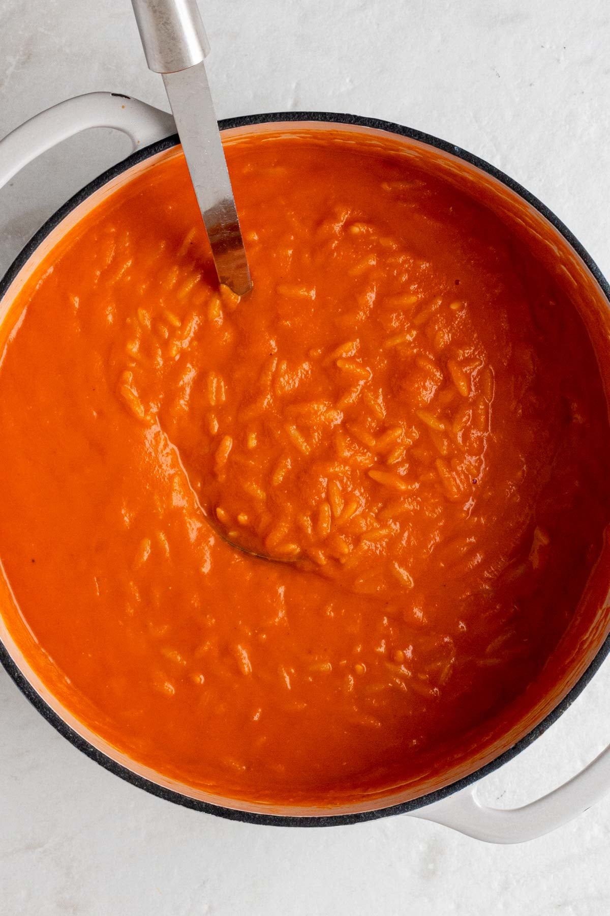 Large beige pot of creamy tomato soup with orzo with a silver ladle on a white background.