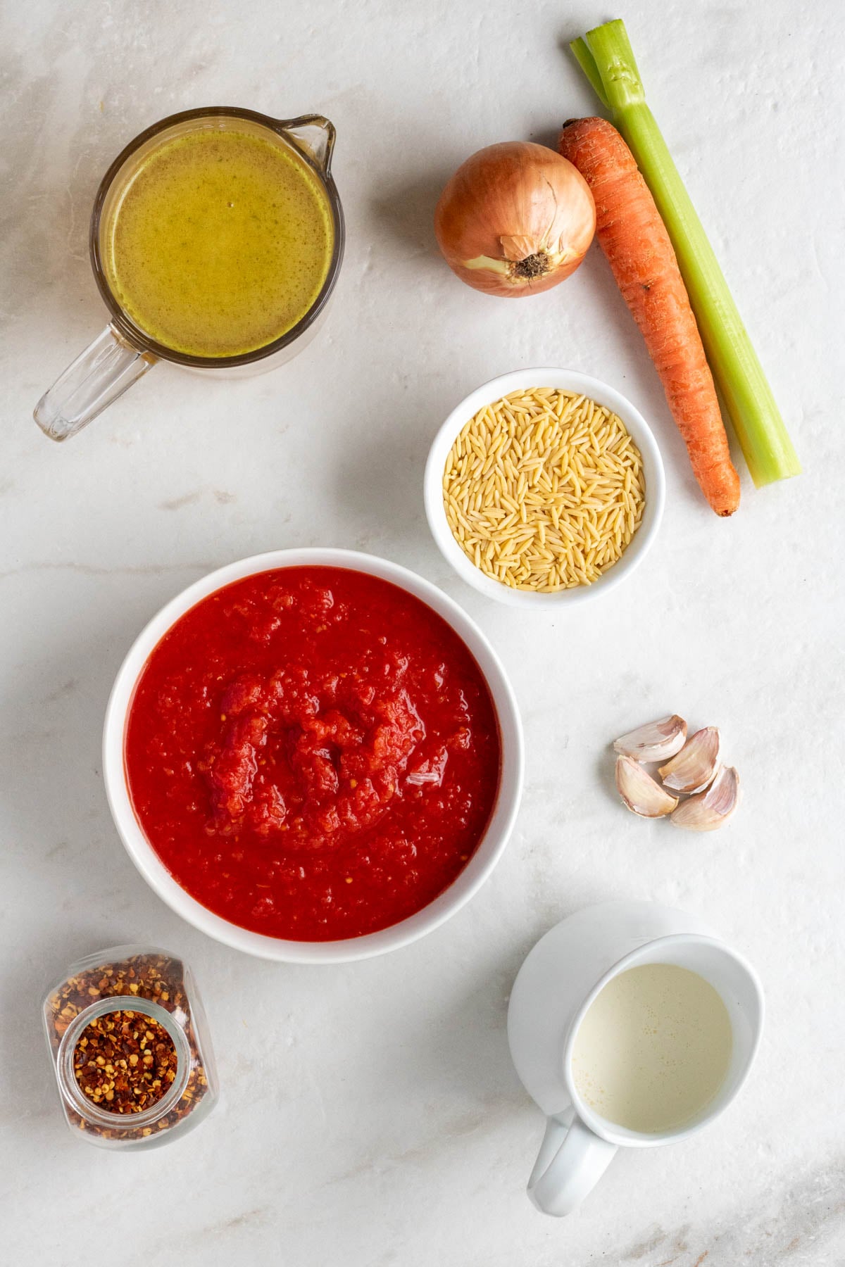 Jar of chicken broth, onion, carrot, celery stick and garlic cloves with a bowl of crushed tomatoes, jar of red pepper flakes, and jug of heavy cream on a white background.