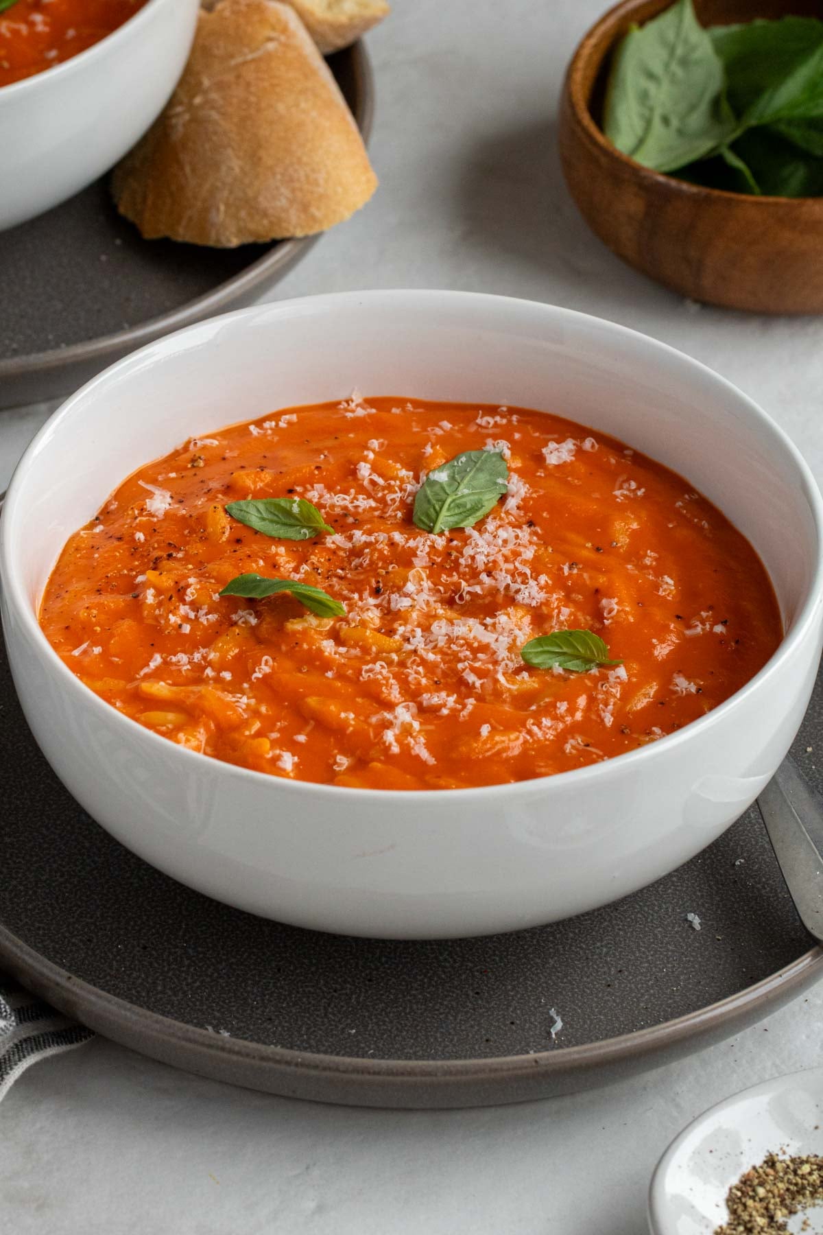 Close up of a white bowl of tomato orzo soup with freshly grated parmesan cheese and basil leaves on top on a dark grey plate on a white background.