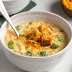 Close up of a bowl of chicken broccoli cheddar soup with croutons and shredded cheese on top on a white background.