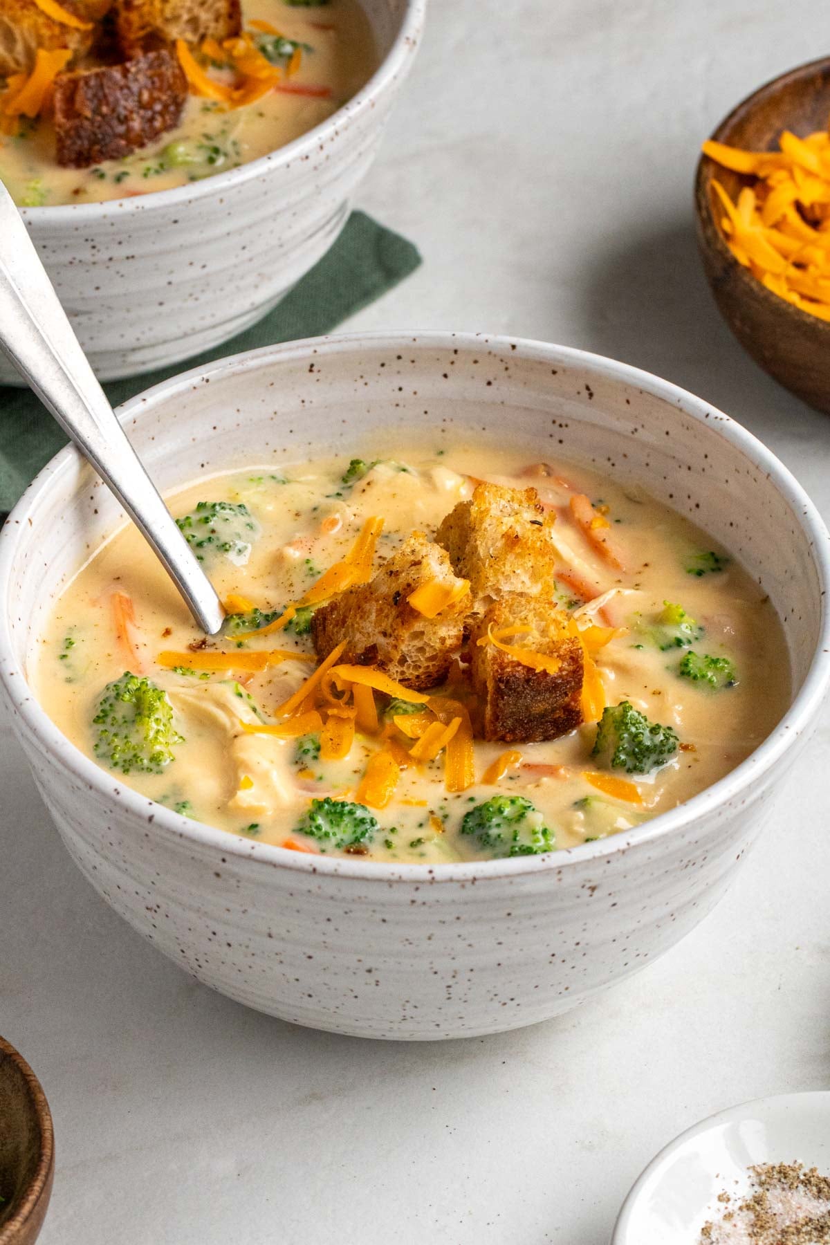 Close up of a bowl of chicken broccoli cheddar soup with croutons and shredded cheese on top on a white background.