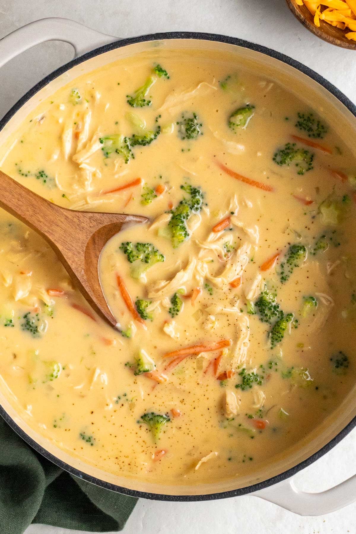 Large pot of broccoli cheddar soup with chicken with a wooden spoon and dark green tea towel on the side on a white background.