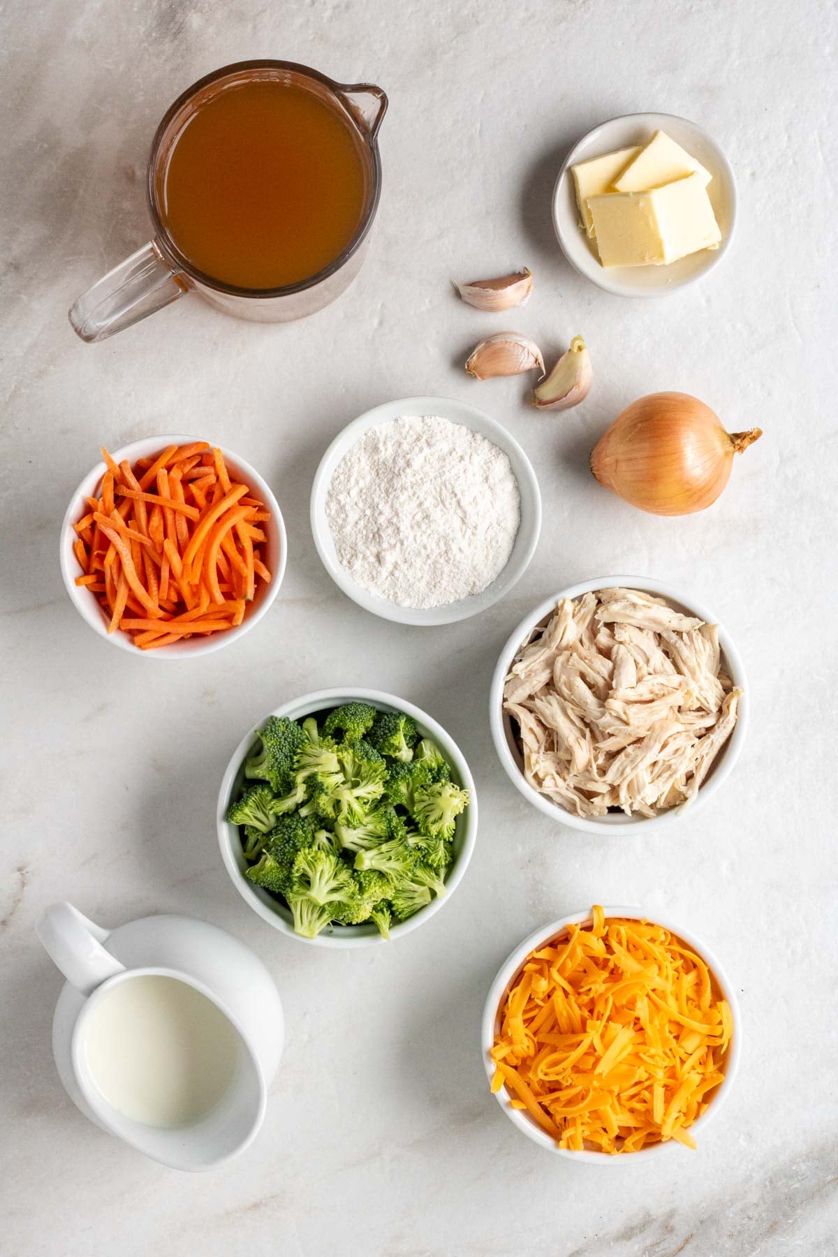 Jug of chicken broth, bowl of butter, garlic cloves, onion, bowl of shredded carrots, bowl of flour, bowl of broccoli florets, bowl of shredded chicken, jug of milk, and bowl of shredded cheddar cheese on a white background.