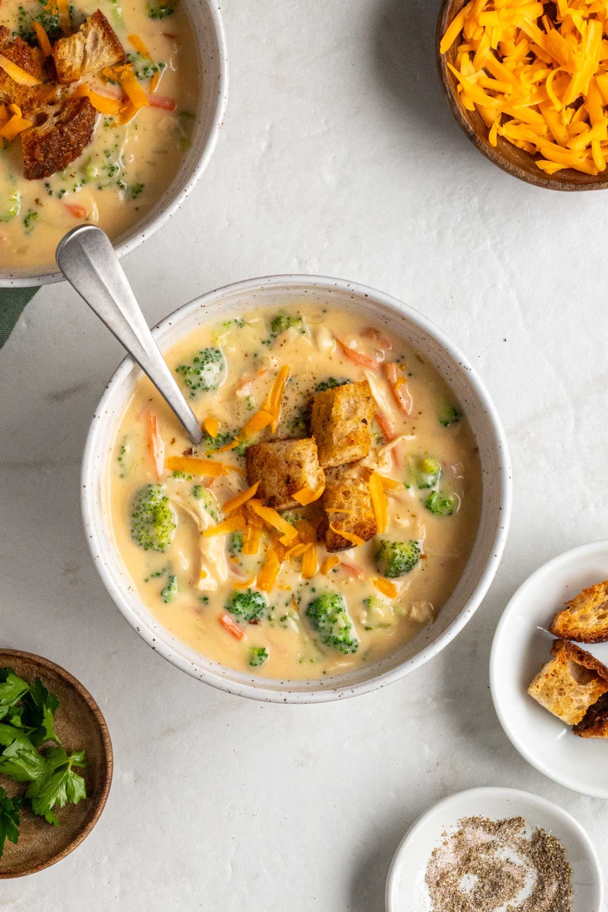 Two white bowls of chicken broccoli cheddar soup with croutons and shredded cheese on top with a wood bowl of fresh parsley and bowls of cheese and croutons on the side on a white background.