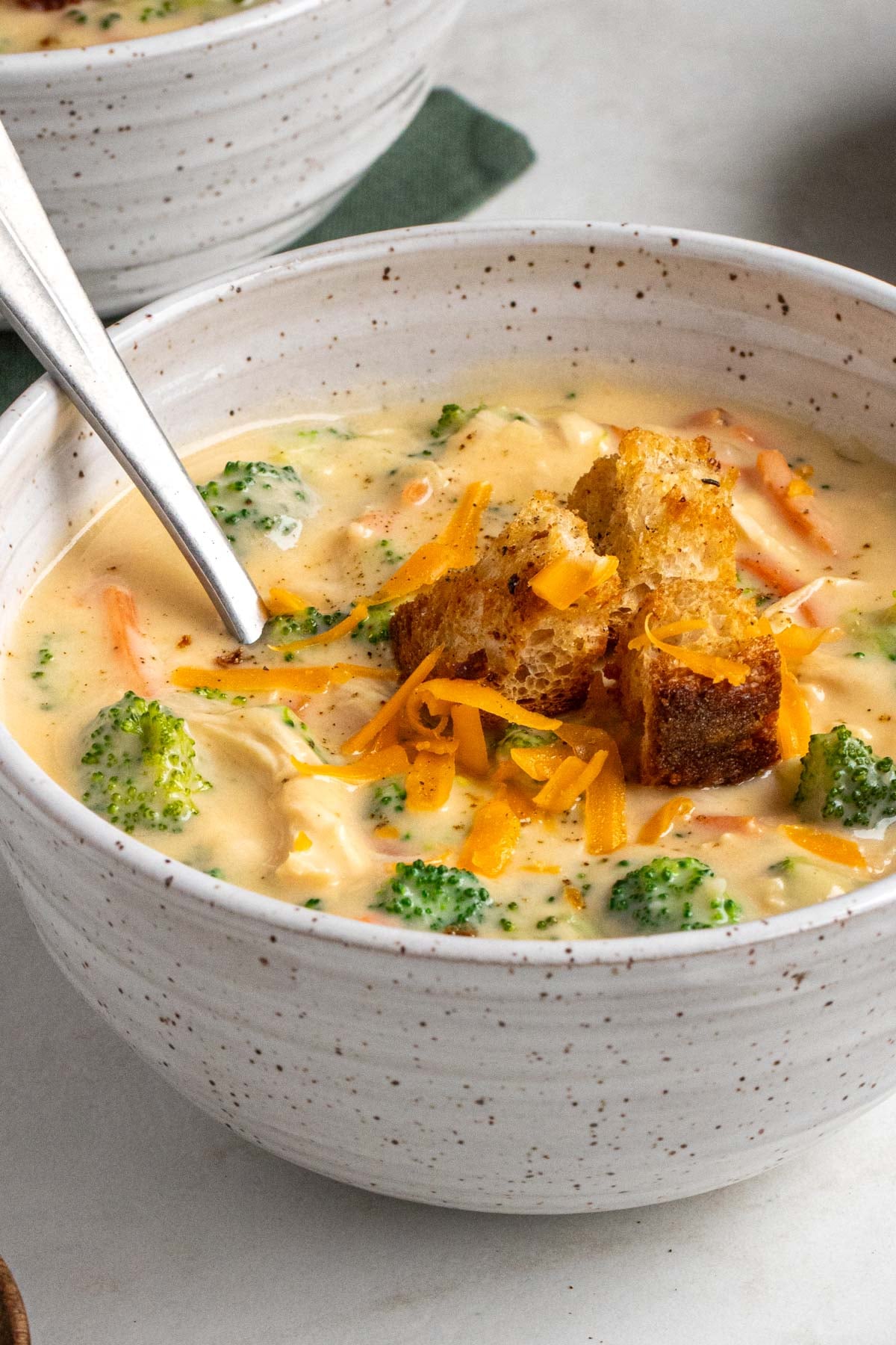 Close up of a chicken broccoli cheese soup in a white speckled bowl with a silver spoon on a white background.