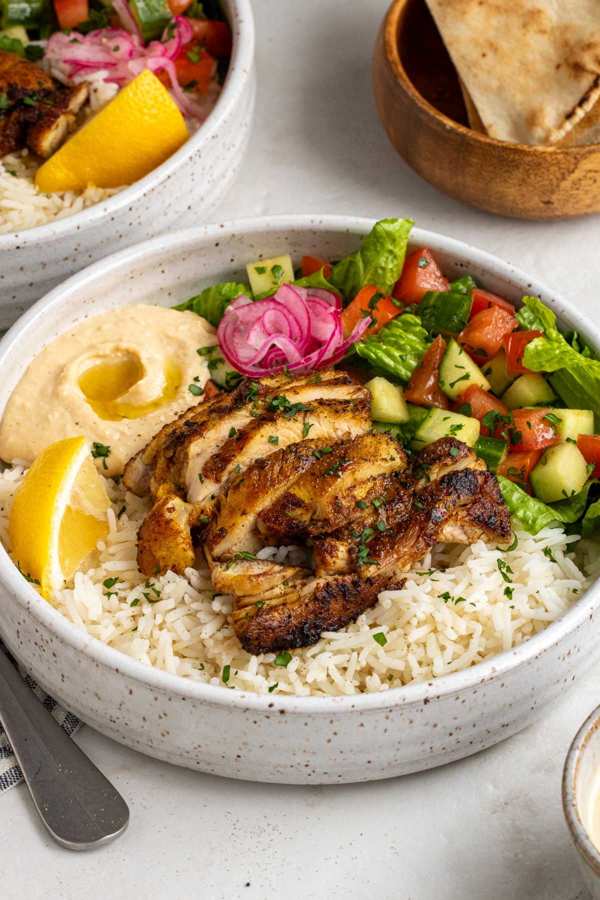 Two chicken shawarma bowls with a wood bowl of pita chips in the background on a white counter.