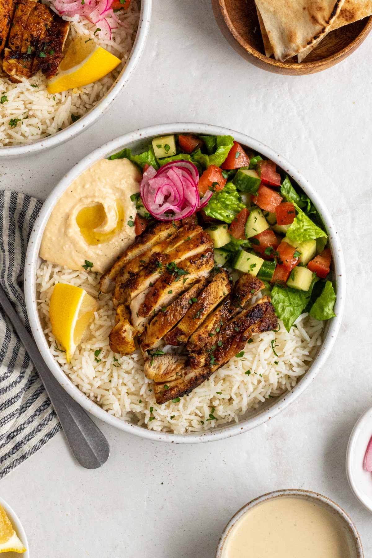 A chicken shawarma bowl with a silver fork and a stripped grey tea towel with sliced pita, tahini sauce, and pickled red onions on the side on a white background.