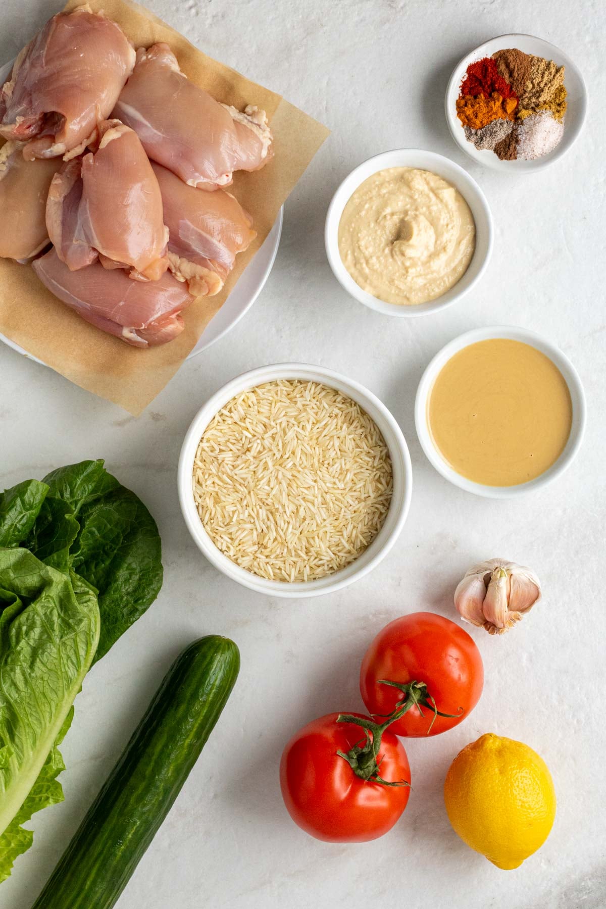 Plate of raw chicken thighs, bowl of shawarma spices, bowl of hummus, bowl of tahini, bowl of white rice, garlic cloves, tomatoes, lemon, cucumber, and romaine lettuce on a white background.