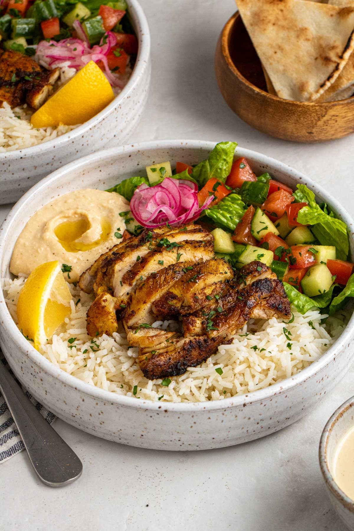 Close up of chicken shawarma bowl with cooked rice, tender chicken, lemon wedge, hummus, tomato cucumber salad, and pickled red onion with chopped parsley on top with garlic tahini sauce on the side on a white background.
