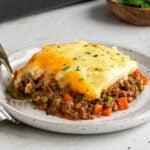 Plate with a slice of shepherd's pie with a gold fork with a wood bowl of fresh parsley and grey casserole dish in the background on a white background.