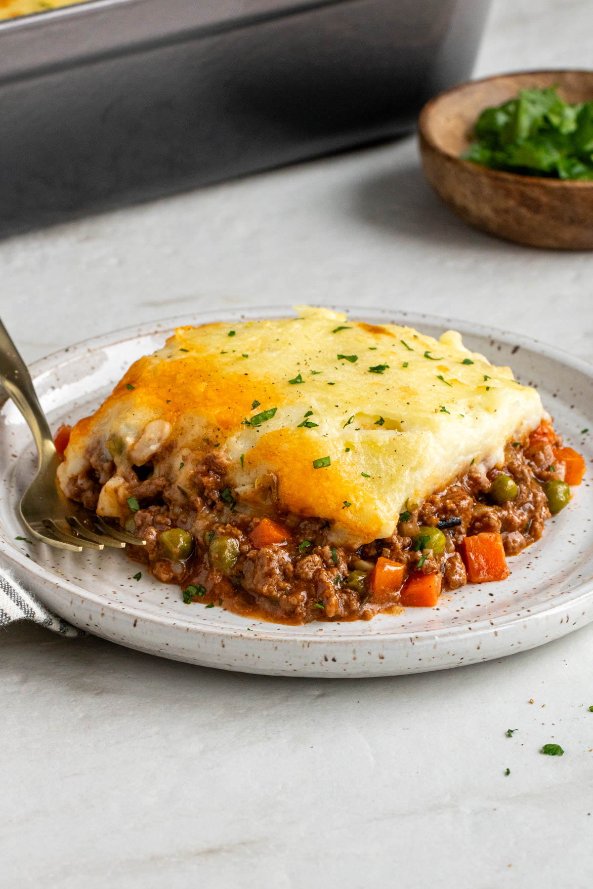 Plate with a slice of shepherd's pie with a gold fork with a wood bowl of fresh parsley and grey casserole dish in the background on a white background.