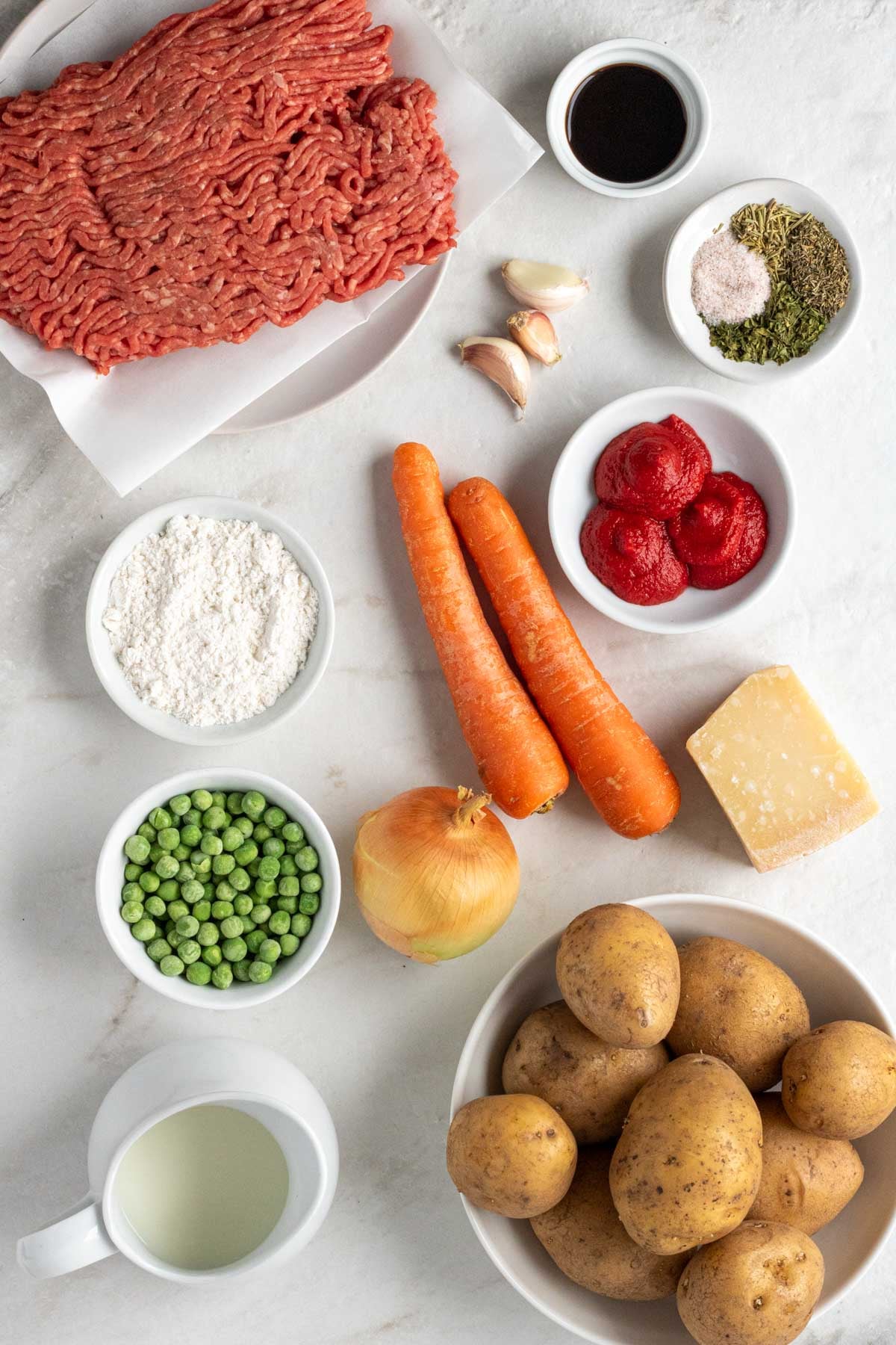 Plate of lean ground beef, bowl of Worcestershire sauce, bowl of dried herbs, bowl of tomato paste, flour, garlic, carrots, onion, parmesan, bowl of frozen pean, jug of milk, and bowl of potatoes on a white background.