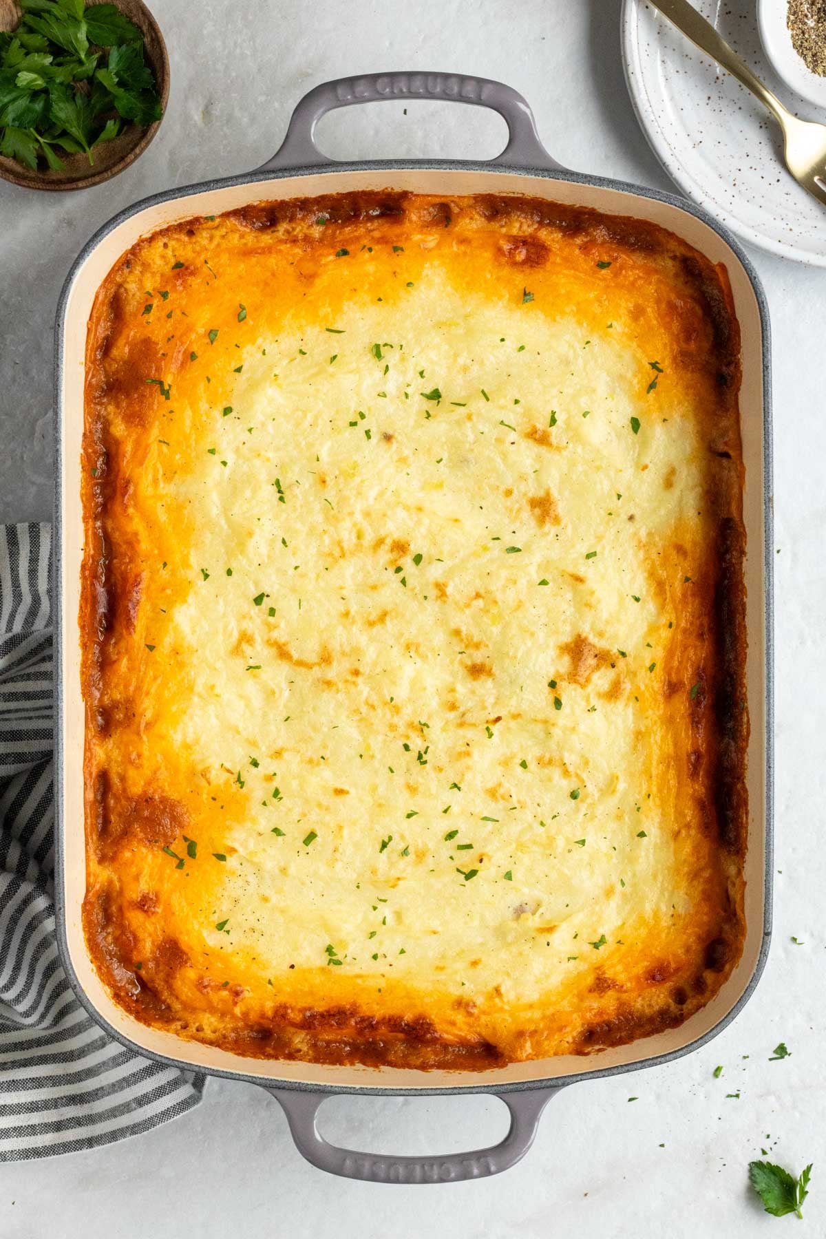 Large baking dish of shepherd's pie with a bowl of fresh parsley, plate, tea towel on the side on a white background.