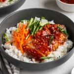 Close up of two spicy gochujang chicken bowls in a dark grey bowl with sliced green onions on top and a small bowl of gochujang sauce in background.