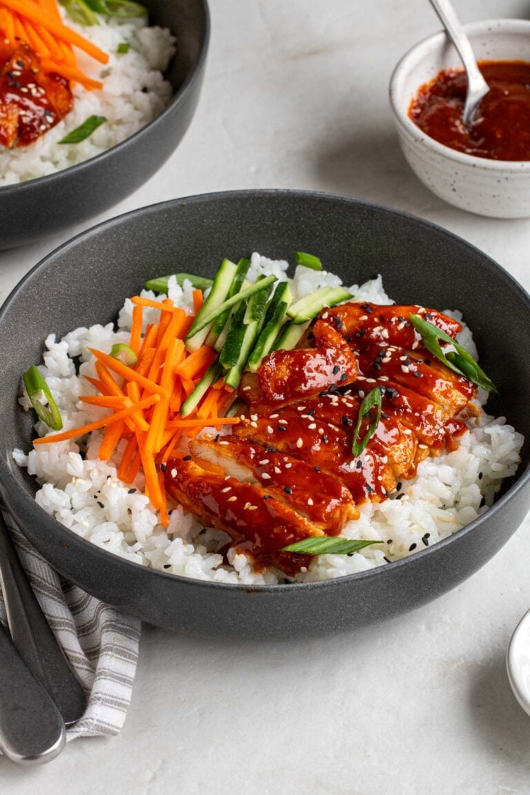 Close up of two spicy gochujang chicken bowls in a dark grey bowl with sliced green onions on top and a small bowl of gochujang sauce in background.