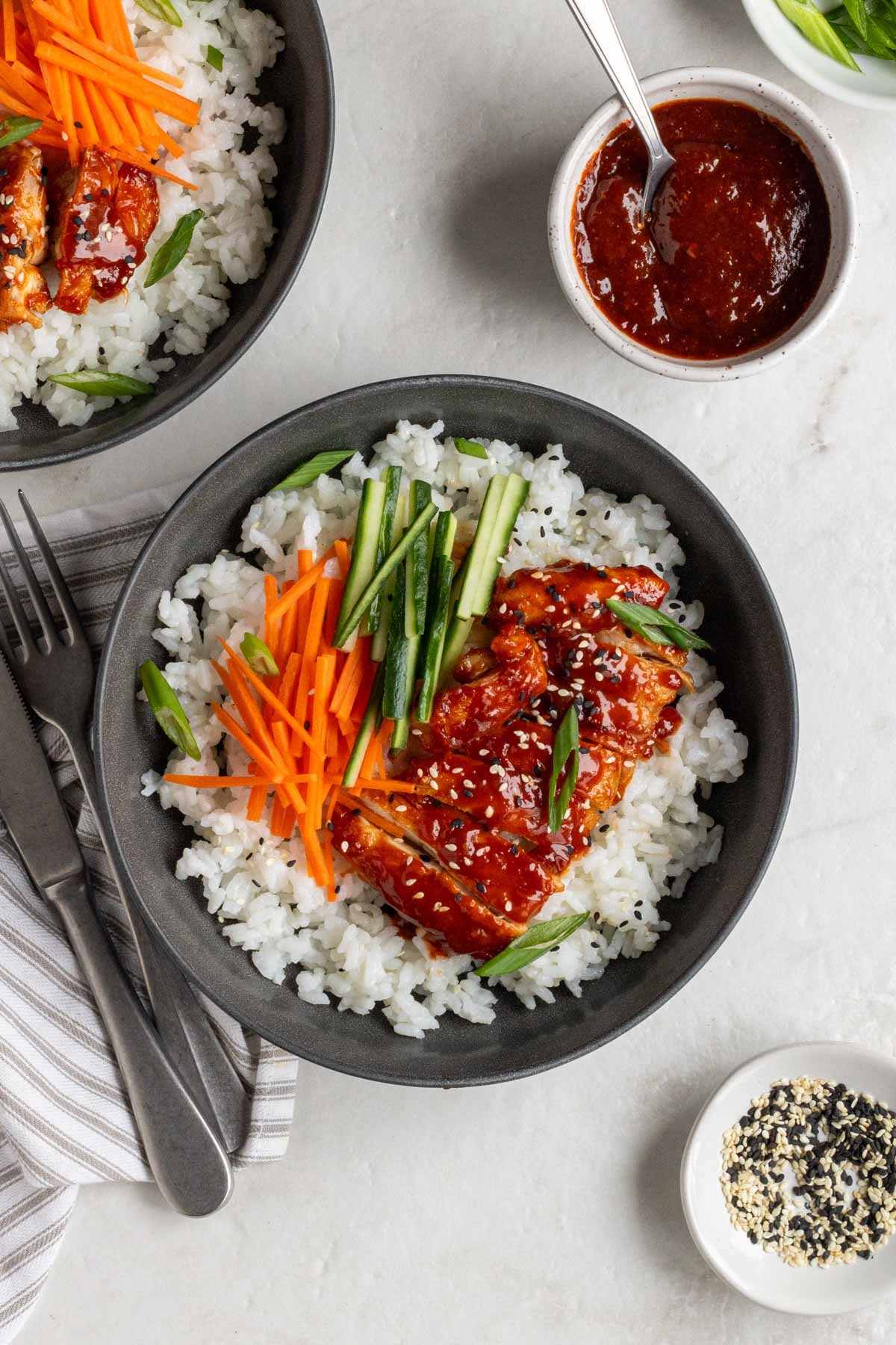 Two gochujang chicken bowls with rice on a white background with a grey and white tea towel, bowl of gochujang sauce, fork and knife, small bowl of black and white sesame seeds on the side.